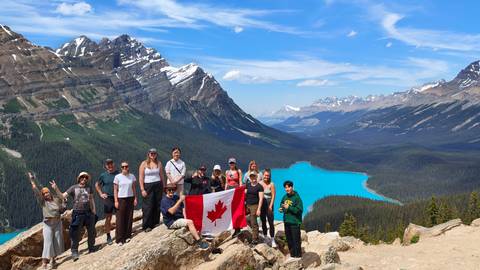 A hiking group waves a Canadian flag atop a rocky viewpoint overlooking turquoise Peyto Lake and the surrounding Rocky Mountains.