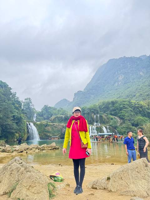Traveller in bright jacket poses before cascading multi-tiered waterfalls amid green mountains.