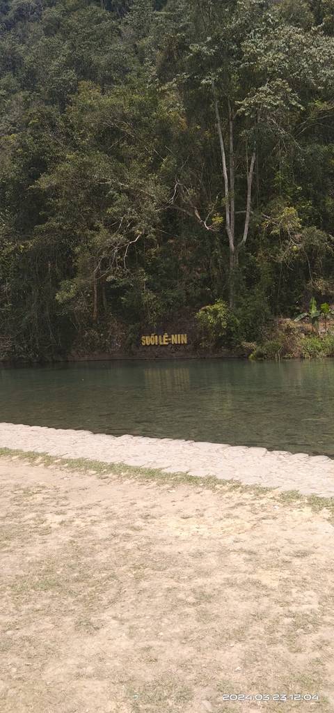 Clear green river beside forested bank with yellow sign reading 'SUỐI LÊ-NIN'.