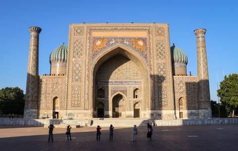 Majestic Sher-Dor Madrasa at Registan Square with visitors photographing the facade in evening light.