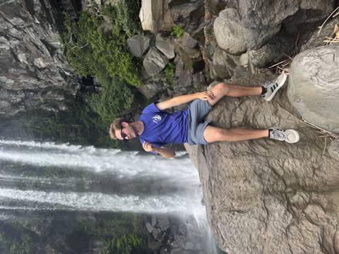 Traveller sitting on a rock eating a snack beside a tall waterfall and lush cliff.