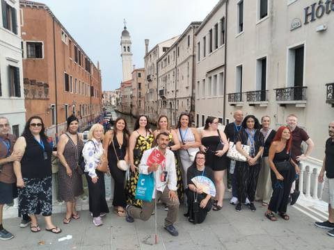 Large tour group posing on a Venetian bridge with narrow canal and bell tower in background.