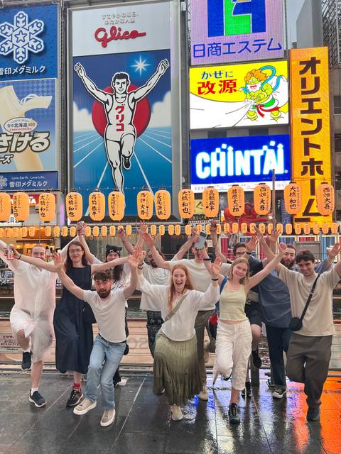 Tour group celebrating by canal in Dotonbori with bright kanji billboards and lanterns.