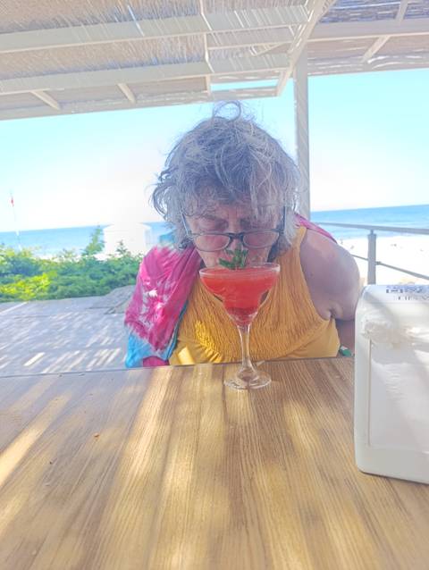 Elderly woman playfully smells a bright red cocktail at an outdoor seaside café.
