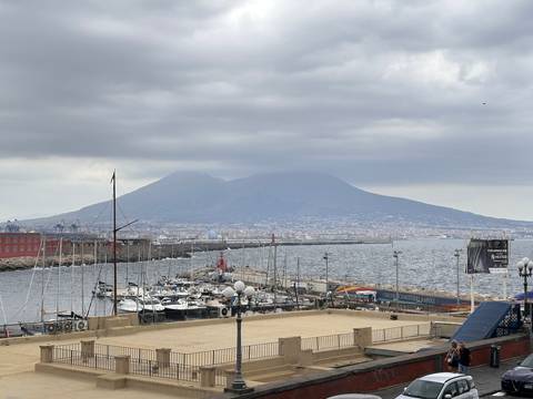 View over a marina and breakwater toward the brooding silhouette of Mount Vesuvius across Naples Bay under gray skies.