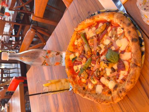 Freshly baked Neapolitan pizza topped with vegetables and mozzarella sits on a wooden table beside iced tea and a glass bottle.