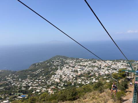 Chairlift cables cut across a high aerial view of Capri island and the endless sea.