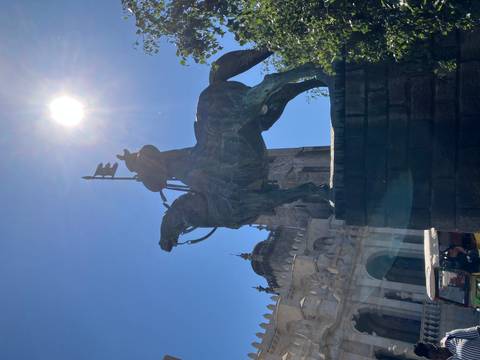 Silhouette of an equestrian statue standing before an old stone cathedral beneath a bright blue sky and sun.