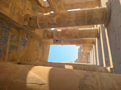 Sunlit sandstone columns with faded painted ceilings inside an ancient Egyptian temple corridor.