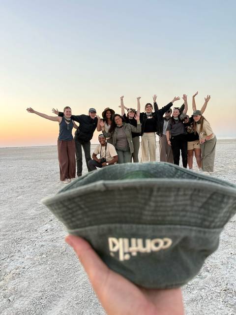 A cheerful tour group poses with arms raised on a vast salt flat at sunset, with a hat brim blurred in the foreground.