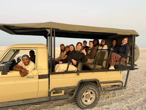 Travelers smile from an open safari truck parked on a flat dusty plain during golden evening light.