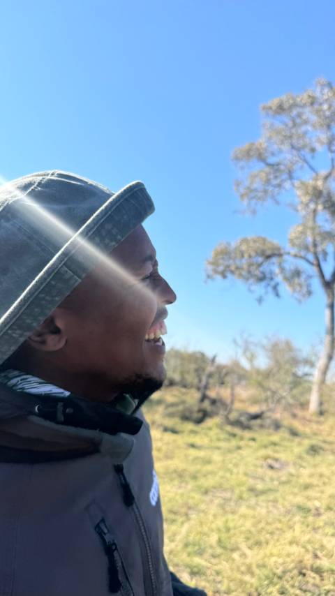 Close-up of a smiling traveler in bright daylight with a sunbeam streaking across the frame and trees behind.