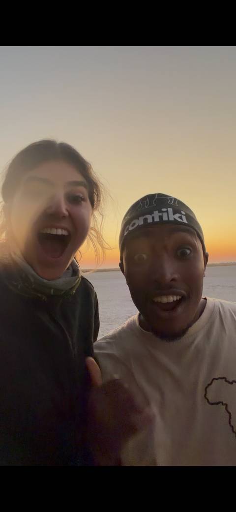 Energetic selfie of two travelers at sunset on a salt flat, faces lit by warm light.