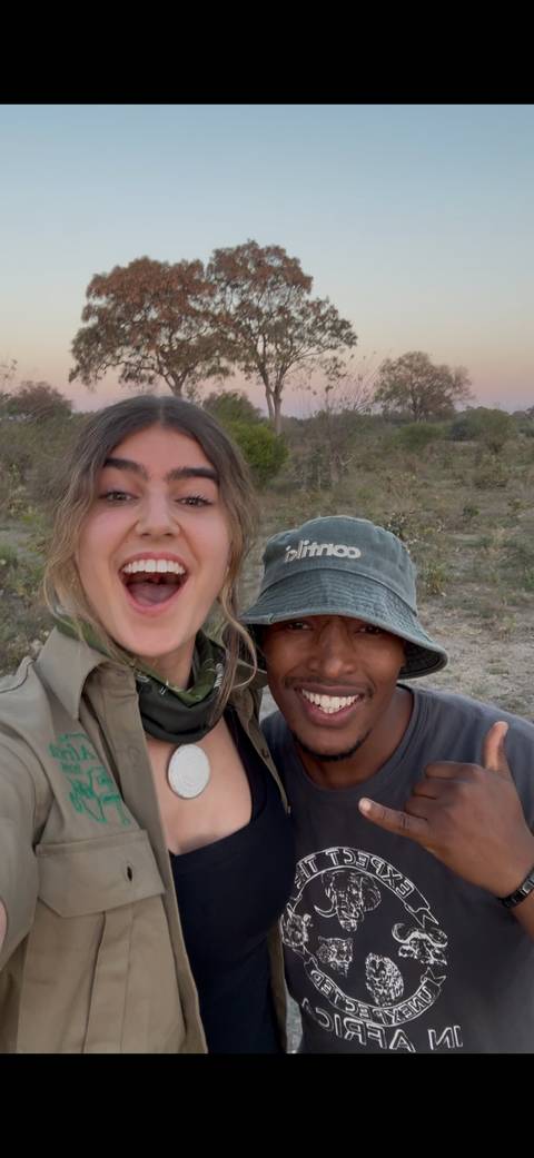 Two smiling travelers pose together outdoors in safari attire against a scrubland backdrop.