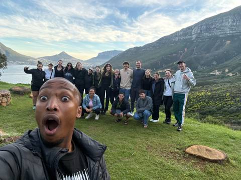 Group posing on grassy hill with dramatic coastal mountains and bay behind; one person making a surprised selfie face