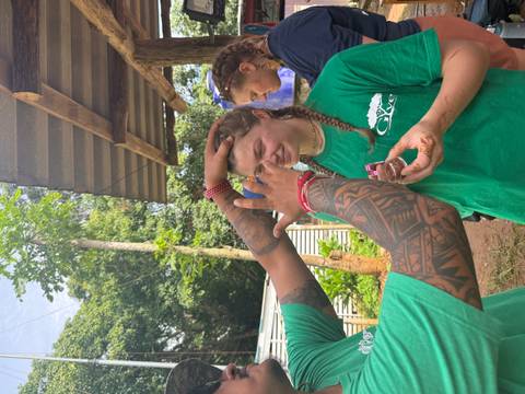 Man with tattooed arm playfully dabs mud on smiling woman’s forehead while others watch in a lush yard.