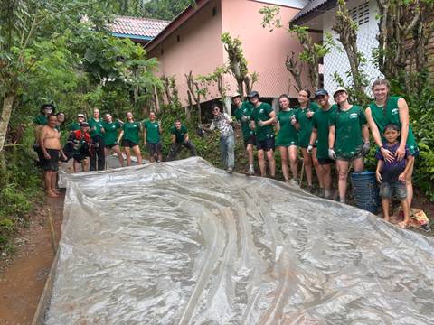 Large group of young volunteers in green shirts standing beside a long plastic sheet on a muddy jungle path.