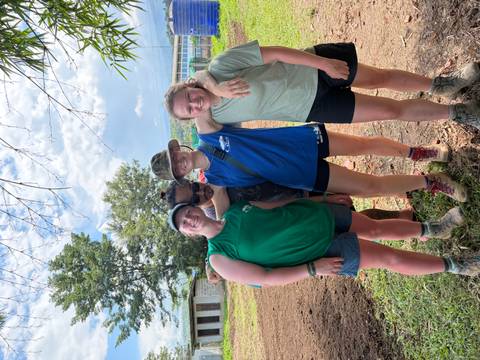 Four muddy volunteers in casual sportswear pose arm-in-arm on a rural worksite with trees and blue sky.