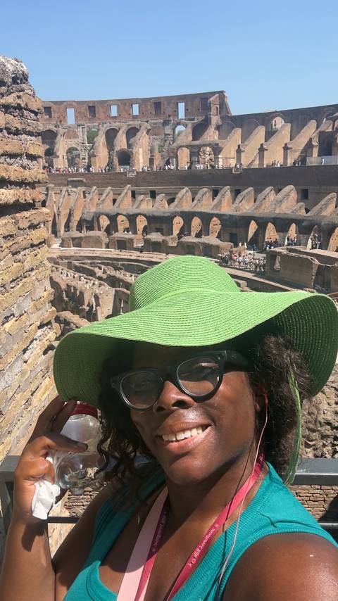Close-up selfie in a large green sunhat with arches and interior tiers of the Colosseum visible behind.