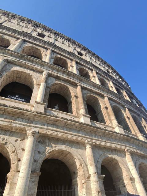 Dramatic upward angle of the stone arches of the Colosseum against a clear blue sky.
