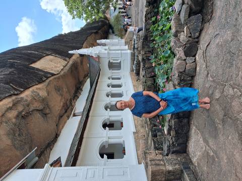 Woman in a blue skirt stands before a white colonnaded temple built beneath a massive overhanging rock.