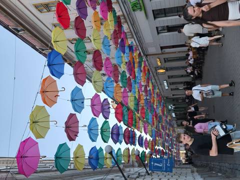 Vibrant pedestrian street covered by a colourful canopy of floating umbrellas with shoppers and cafés below.
