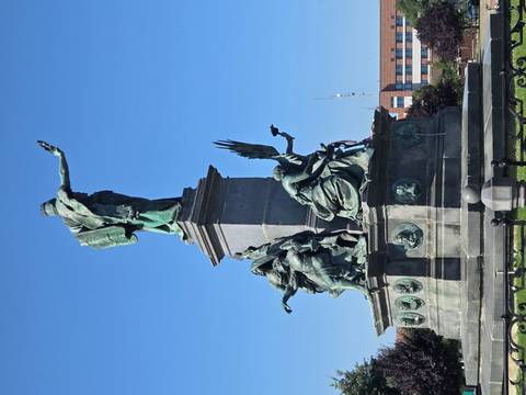 Bronze statue ensemble on a tall plinth depicting historical figures against a clear blue sky in a city square.