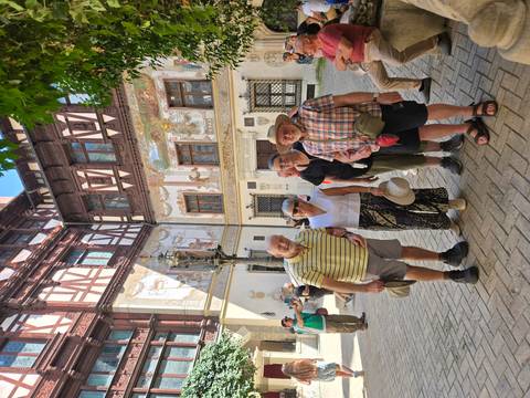 Group of senior travellers pose in a historic courtyard adorned with frescoed walls and timber framing.