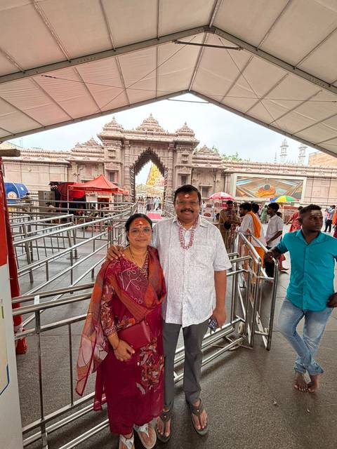 Smiling couple with tilak marks pose at the entrance of a Hindu temple complex with metal queue rails.