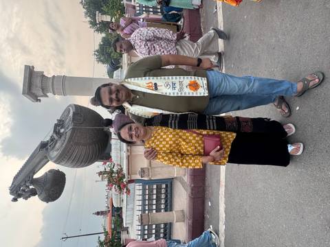 Smiling couple in front of a giant veena sculpture and stone pillar on a busy street.