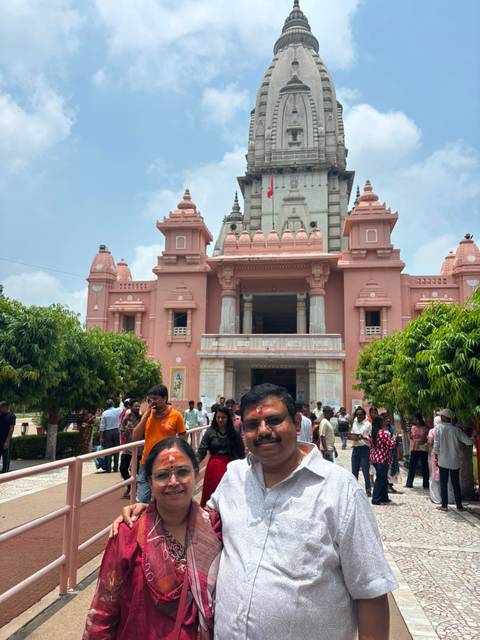 Crowds gather outside a pink-coloured Hindu temple with trees and bright mid-day light.