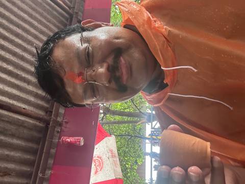 A middle-aged man wearing an orange rain poncho and tilak smiles while holding a small clay cup of tea inside a covered stall.