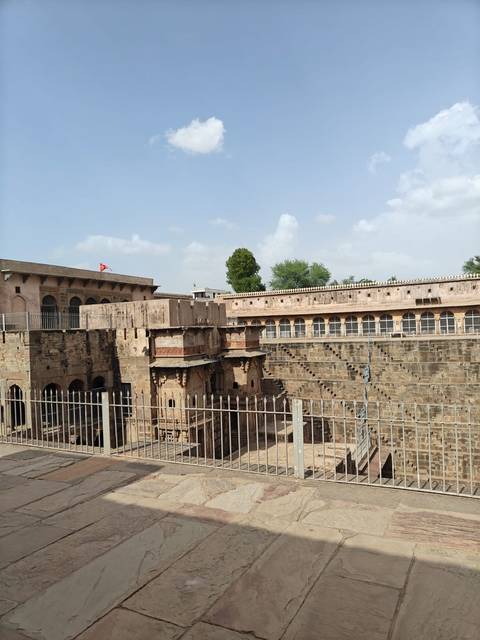View of Chand Baori stepwell and surrounding sandstone structures behind a metal railing on a sunny day.