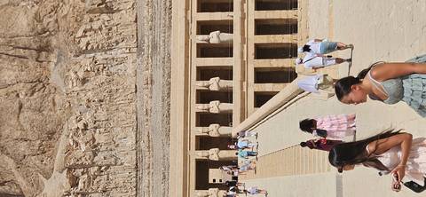 Visitors walk up the broad ramp toward the colonnaded facade of an ancient Egyptian temple carved into a cliff.