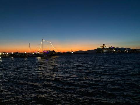 Colorful sunset over a harbor with sailboats and a lit cruise ship silhouetted against the horizon