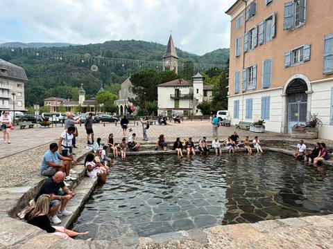 Large tour group relaxing around a shallow stone pool in a quaint European village square