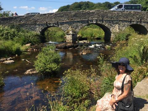 Woman in sunhat stands beside an old triple-arched stone bridge over a clear stream in the countryside