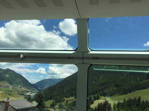 View from inside a panoramic train carriage showing blue skies, clouds and alpine valleys through large windows