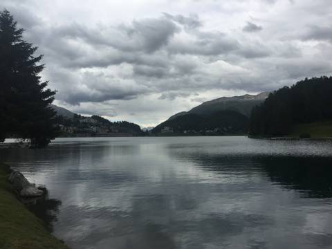 Moody lake surrounded by dark evergreen forest and distant alpine village under heavy clouds