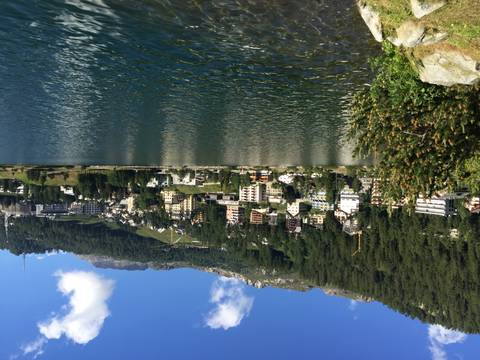 Lakeside view of alpine resort town backed by forested slopes and rocky peaks under blue sky