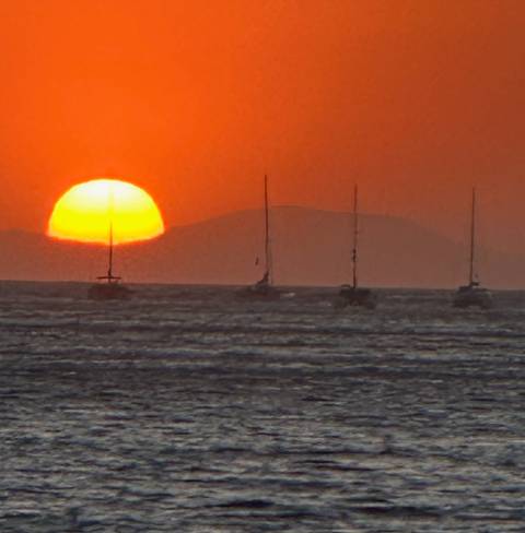 Brilliant orange sunset over the sea with silhouettes of several sailboats and a distant island.