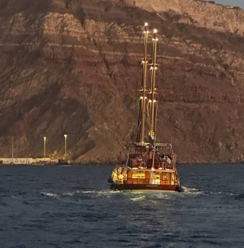Traditional wooden sailboat moving toward rugged volcanic cliffs at dusk.