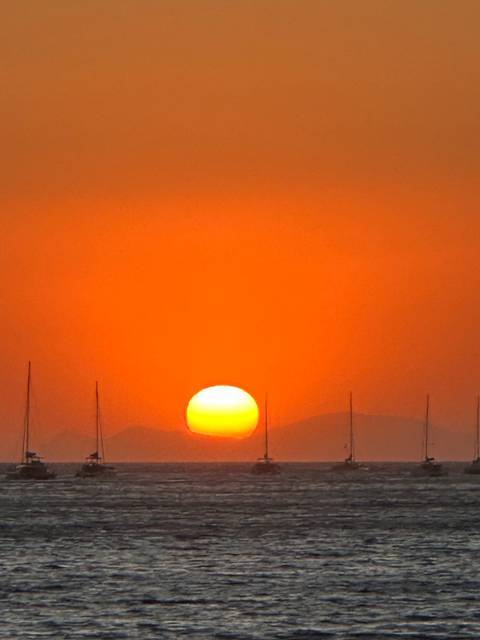Intense orange sky with the sun sinking below distant hills and faint sail-masts.