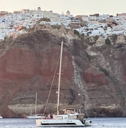 Sailboat mast set against red volcanic cliffs topped with white buildings of Santorini.