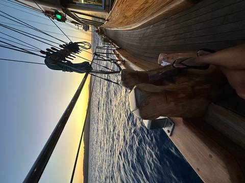 Relaxed view from a wooden sailboat deck at dusk with rigging lines and a passenger’s feet.