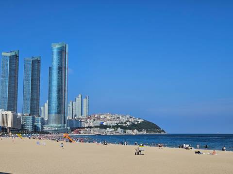 Busy beach scene with tall modern skyscrapers rising behind crowded sand and blue sea.