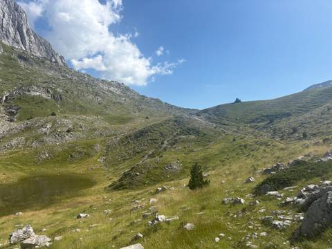 Grassy alpine hollow with a small pond nestles amid rocky slopes under a bright sky.
