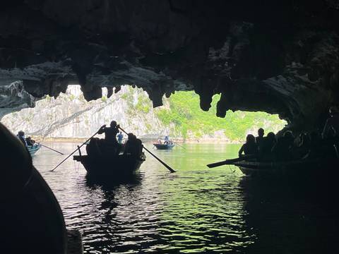 Silhouetted rowboats glide through a limestone cave opening onto bright green cliffs and calm waters in Ha Long Bay.