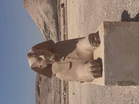 A sandstone sphinx statue sits on a plinth with barren hills and blue sky behind.