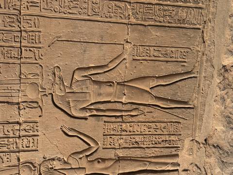 Close-up relief carving of an Egyptian god with hieroglyphics on a sandstone wall lit by warm sunlight.